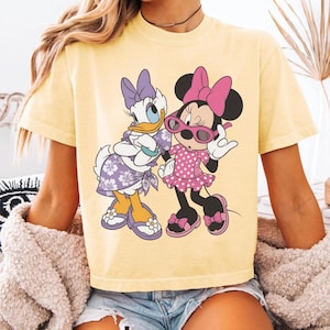 Puede incluir: Una camiseta de color amarillo p&aacute;lido con un estampado gr&aacute;fico de Daisy Duck y Minnie Mouse. Daisy lleva un vestido morado con flores y un lazo, mientras que Minnie lleva un vestido rosa de lunares, un lazo rosa y gafas de sol rosas. Ambas llevan sandalias.