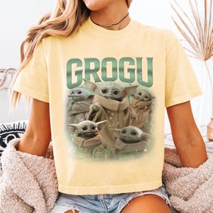 May include: A light yellow t-shirt featuring the word "GROGU" in green block letters above a graphic of Grogu from The Mandalorian. The graphic shows Grogu in various poses. The shirt has short sleeves and a crew neck.