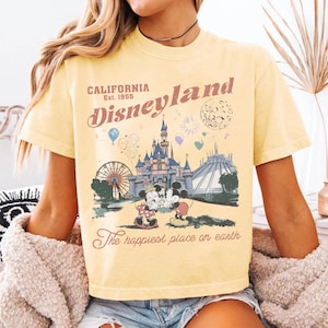 Op de afbeelding: Lichtgeel T-shirt met een Disneyland-afbeelding met Mickey en Minnie Mouse, een kasteel en de tekst "California Est. 1955". Het shirt heeft ook de tekst "The happiest place on earth."