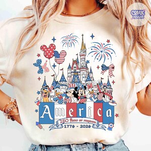 May include: A cream-colored t-shirt featuring a graphic of a castle, fireworks, and cartoon characters in patriotic red, white, and blue. The text reads America, 250 Years of Freedom, and 1776-2026, with Mickey-shaped balloons and stars.