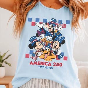 May include: A light blue sleeveless tank top featuring a graphic of Disney characters including Mickey Mouse, Minnie Mouse, Donald Duck, Daisy Duck, Goofy, and Pluto wearing patriotic hats. The text reads AMERICA 250 and 1776-2026.