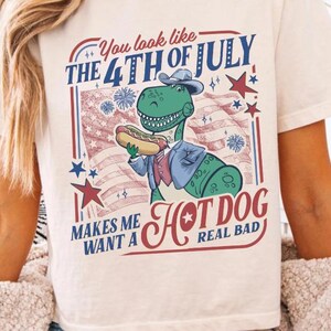 May include: Cream-colored t-shirt featuring a graphic of a dinosaur holding a hot dog, with text that reads "You look like the 4th of July makes me want a hot dog real bad." The design includes red, white, and blue elements.