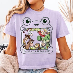 May include: A light purple t-shirt featuring a graphic of a frog outline containing various animated characters from the Toy Story franchise. The text "YOU'VE GOT A FRIEND IN ME" is printed in black capital letters below the character illustration.