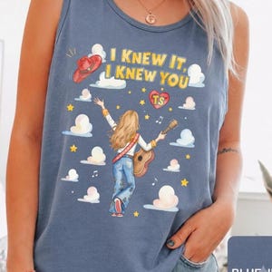 May include: A blue denim-colored tank top featuring a graphic of a person with long blonde hair playing an acoustic guitar. The text reads I KNEW IT, I KNEW YOU, accompanied by a red cowboy hat, a heart with TS, yellow stars, and white clouds.