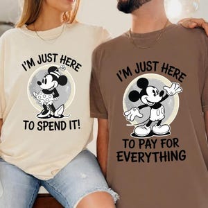May include: Two t-shirts, one beige and one brown, featuring Mickey Mouse designs. The beige shirt has Minnie Mouse with the text "I'M JUST HERE TO SPEND IT!". The brown shirt has Mickey Mouse with the text "I'M JUST HERE TO PAY FOR EVERYTHING".