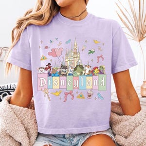 May include: A light purple t-shirt featuring a Disneyland graphic. The graphic includes a castle, various cartoon characters, and the word "Disneyland" in a colorful font. The shirt is short-sleeved.