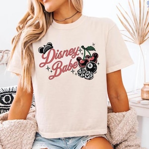 May include: A cream-coloured t-shirt featuring the text "Disney Babe" in pink script, alongside a graphic of cherries, an eight ball, and dice. The shirt has short sleeves and a crew neck.