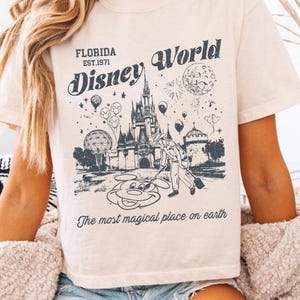 K&ouml;nnte beinhalten: Cremefarbenes T-Shirt mit einer Grafik von Disney World in Florida. Das Design zeigt ein Schloss, Mickey Mouse und den Text "Florida Est. 1971 Disney World" und "The most magical place on earth."