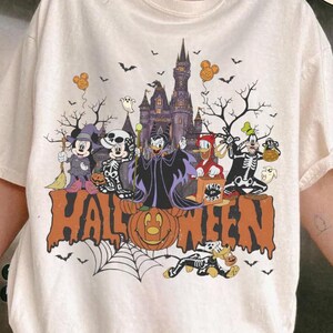 May include: An ivory t-shirt featuring a graphic print of Disney characters in Halloween costumes in front of a castle. The text reads HALLOWEEN, COMFORT COLORS IVORY, and COMIC DESIGNS. The design includes Mickey, Minnie, Goofy, and Donald.
