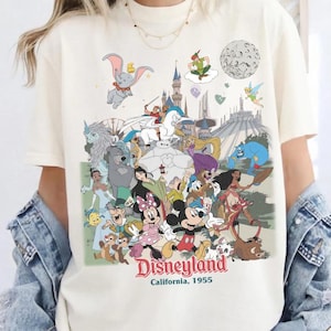 Puede incluir: Camiseta de color marfil con un gr&aacute;fico colorido de Disneyland. El dise&ntilde;o incluye varios personajes de Disney, un castillo y las palabras "Disneyland California, 1955". La camiseta es de Comfort Colors y tiene un logotipo de Comic Designs.