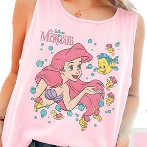 May include: A light pink sleeveless tank top featuring a graphic print of Ariel from The Little Mermaid with her long pink hair, a purple seashell top, and the yellow fish Flounder surrounded by blue bubbles and small yellow seahorses.