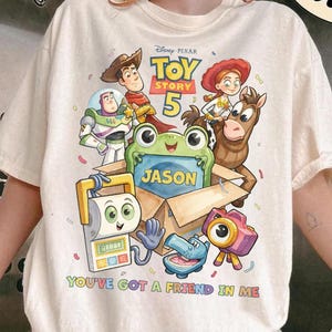 May include: An ivory t-shirt featuring a Toy Story 5 graphic with characters including Woody, Buzz Lightyear, Jessie, and Bullseye. A green frog sits in a cardboard box labeled Jason. The text reads Toy Story 5 and You've Got A Friend In Me.