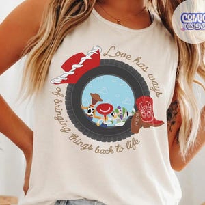 May include: An ivory sleeveless tank top featuring a graphic print of a black tire framing a blue sky scene with cartoon characters. The design includes a red cowboy hat, a red cowboy boot, and the text Love has ways of bringing things back to life.
