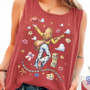 May include: A brick red sleeveless tank top featuring a graphic illustration of a person with long hair playing a guitar, wearing cow print bell-bottom pants and a hat. The design includes flowers, clouds, stars, a bow, and blocks with the text BTS.
