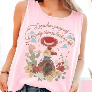 Op de afbeelding: Een lichtroze mouwloze tanktop met een grafische print van een cowgirl met een rode hoed, zittend op een paard te midden van cactussen en bloemen. De tekst luidt, Love has ways of bringing things back to life, met muzieknoten en wolken.