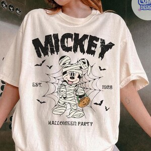 May include: A cream-colored short-sleeve t-shirt featuring a black graphic of Mickey Mouse wrapped as a mummy inside a spider web. The text reads MICKEY, EST. 1928, and HALLOWEEN PARTY. Small black bats are scattered around the central design.