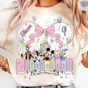 Op de afbeelding: Cr&egrave;mekleurig T-shirt met een kleurrijke Disney-afbeelding. Het toont personages, een roze strik en de tekst "OliviaLand". Het ontwerp bevat het nummer 4, vuurwerk en de zin "The happiest birthday on Earth".
