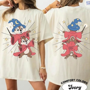 Puede incluir: Camiseta de color marfil con un dise&ntilde;o de mago de dibujos animados en la parte delantera y trasera. El dise&ntilde;o incluye dos ardillas listadas con t&uacute;nicas rojas y sombreros de mago azules, sosteniendo varitas. La camiseta tiene las palabras "COMFORT COLORS" e "Ivory".