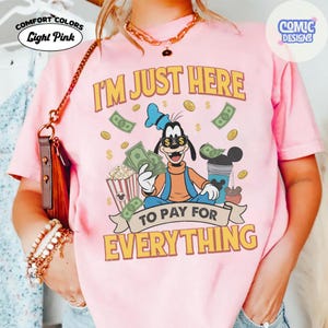 May include: Light pink Comfort Colors t-shirt with the text "I'M JUST HERE TO PAY FOR EVERYTHING". The graphic features a cartoon character holding money and popcorn, with dollar signs and coins.