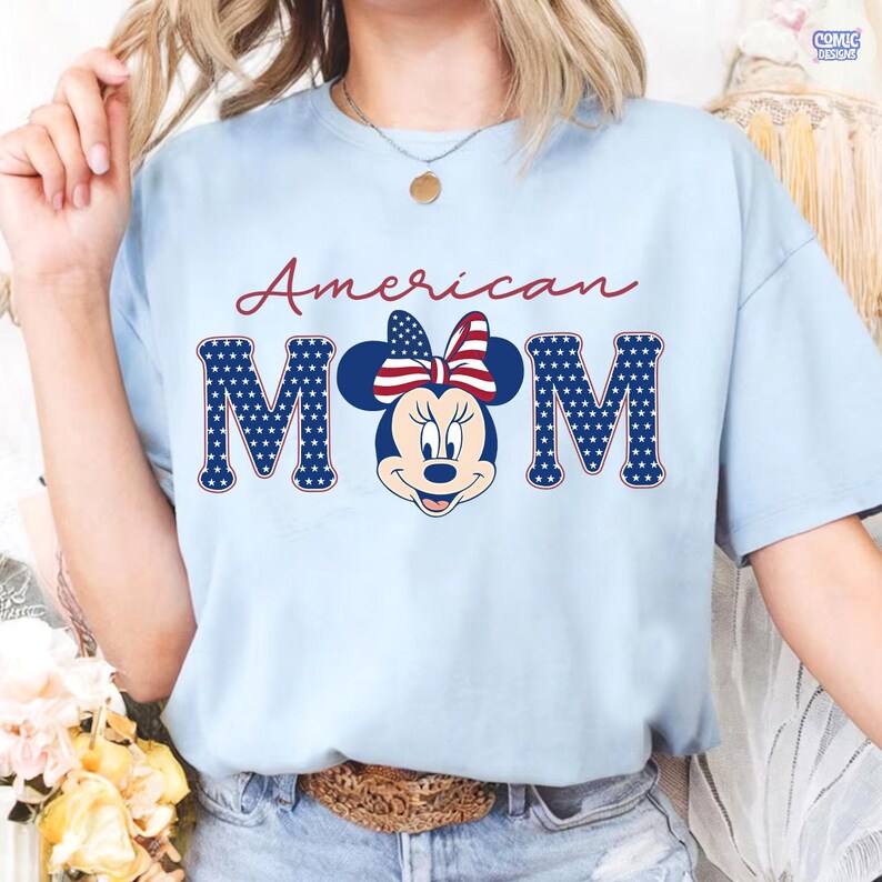 May include: Light blue t-shirt with the words "American Mom" in red script. The letters "MOM" are in blue with white stars, featuring a Minnie Mouse graphic with a patriotic bow.