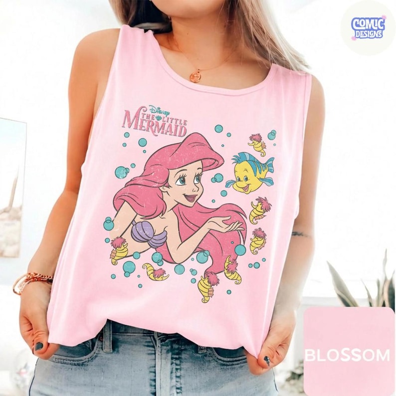 May include: A light pink sleeveless tank top featuring a graphic print of Ariel from The Little Mermaid with her long pink hair, a purple seashell top, and the yellow fish Flounder surrounded by blue bubbles and small yellow seahorses.