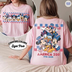May include: Light pink t-shirt featuring a patriotic design with cartoon characters. The front displays a row of characters within a red and blue checkered border. The back features a larger group of characters, the American flag, and the text "AMERICA 250 1776-2026."