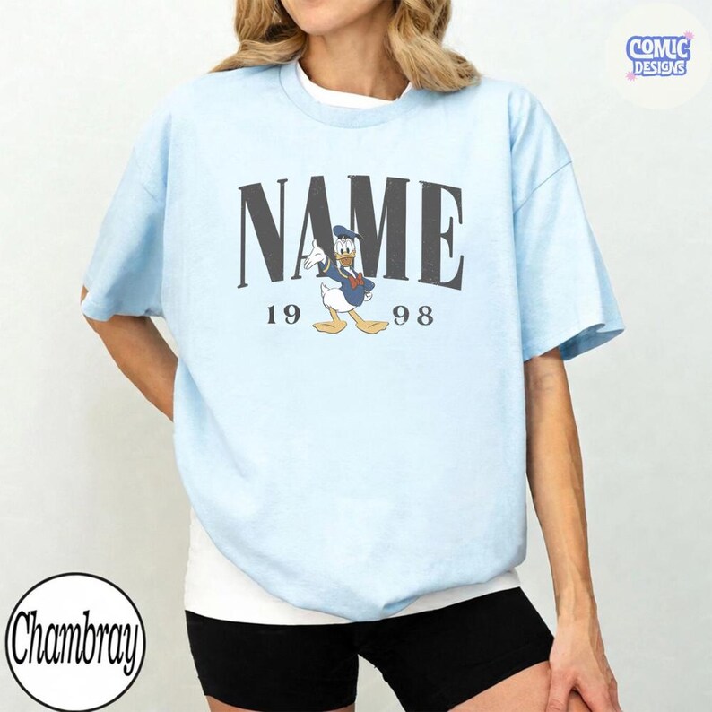 K&ouml;nnte beinhalten: Hellblaues T-Shirt mit dem Wort "NAME" in dunkelgrauen Blockbuchstaben &uuml;ber einer Cartoon-Ente in Matrosenanzug. Die Zahlen "19" und "98" flankieren die Ente. Das Shirt hat kurze &Auml;rmel und einen Rundhalsausschnitt.