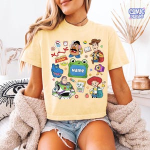 May include: A pale yellow t-shirt featuring a colorful graphic print of various cartoon characters, including a space ranger, a cowgirl, a cowboy, a potato-shaped figure, and a green frog-shaped frame with the word Name printed inside.