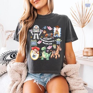 May include: A charcoal gray short-sleeve t-shirt featuring a graphic print of various Toy Story characters including Buzz Lightyear, Woody, Jessie, Rex, Slinky Dog, and Bullseye, with the text "You've Got A Friend In Me" centered among the illustrations.