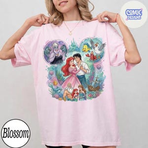 May include: A light pink t-shirt featuring a large graphic print of characters from The Little Mermaid arranged within a Mickey Mouse ear silhouette. The design includes Ariel, Eric, Ursula, King Triton, Flounder, Sebastian, and a ship. The text Comic Designs appears in the top right corner.