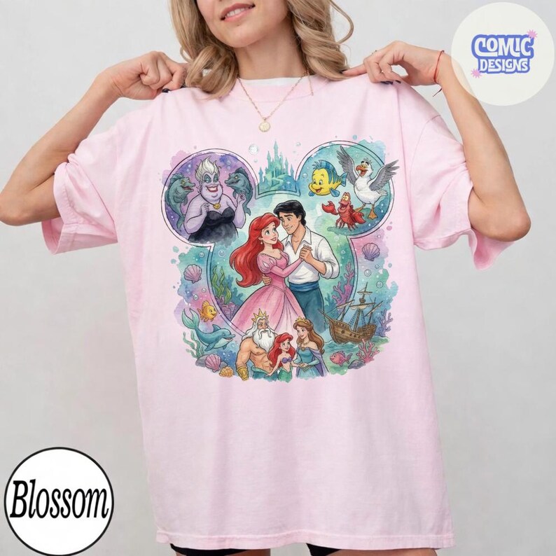 May include: A light pink t-shirt featuring a large graphic print of characters from The Little Mermaid arranged within a Mickey Mouse ear silhouette. The design includes Ariel, Eric, Ursula, King Triton, Flounder, Sebastian, and a ship. The text Comic Designs appears in the top right corner.
