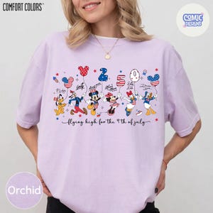 May include: Orchid-colored t-shirt with a graphic featuring Disney characters holding balloons shaped like Mickey Mouse heads and the number "250". The text "flying high for the 4th of July" is below the characters. The shirt has the "Comfort Colors" logo.