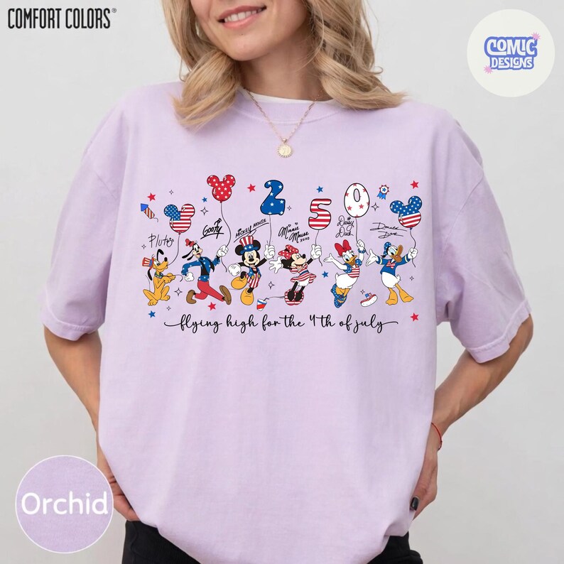 May include: Orchid-colored t-shirt with a graphic featuring Disney characters holding balloons shaped like Mickey Mouse heads and the number "250". The text "flying high for the 4th of July" is below the characters. The shirt has the "Comfort Colors" logo.