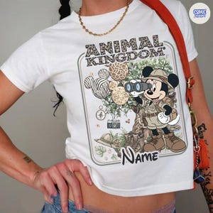 May include: White t-shirt featuring a graphic of Mickey Mouse in safari attire, holding binoculars. The design includes the text "ANIMAL KINGDOM" in a leopard print font, along with animal-themed Mickey Mouse head shapes. A name can be added below the graphic.