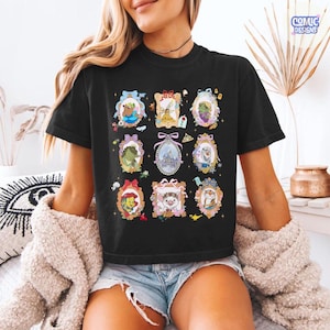 Puede incluir: Camiseta negra con retratos enmarcados de personajes de dibujos animados. Los retratos est&aacute;n en marcos ornamentados con lazos y detalles florales. La camiseta tiene el texto "COMIC DESIGNS" en la esquina superior derecha.
