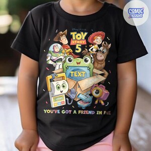 May include: A black short-sleeved t-shirt featuring a Toy Story 5 graphic with characters including Woody, Buzz Lightyear, Jessie, and Bullseye. A green frog sits in a cardboard box with the word TEXT, and the phrase YOU'VE GOT A FRIEND IN ME.