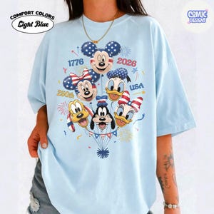 以下が含まれることがあります： ライトブルーのComfort Colors Tシャツ。ミッキーマウス、ミニーマウス、ドナルドダック、デイジーダック、グーフィー、プルートが風船になったグラフィックデザイン。デザインには「1776」、「2026」、「250th」、「USA」の文字が含まれています。