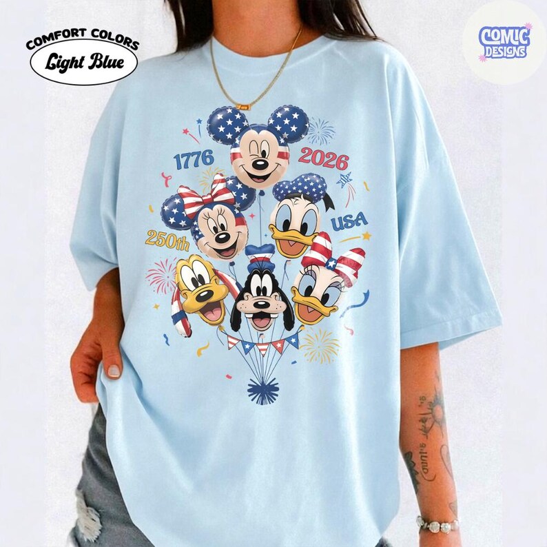 以下が含まれることがあります： ライトブルーのComfort Colors Tシャツ。ミッキーマウス、ミニーマウス、ドナルドダック、デイジーダック、グーフィー、プルートが風船になったグラフィックデザイン。デザインには「1776」、「2026」、「250th」、「USA」の文字が含まれています。