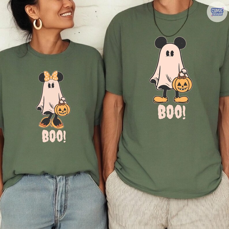 May include: Two olive green t-shirts with a Halloween-themed graphic. The graphic features a cartoon ghost with Mickey Mouse ears holding a jack-o'-lantern and the word "BOO!". One shirt has a bow on the ghost's head.