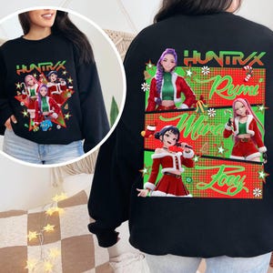 Puede incluir: Sudadera negra con un gráfico festivo de personajes de estilo anime con atuendos navideños. La parte delantera muestra el texto "HUNTRAX" y un grupo de personajes. La parte trasera tiene el texto "HUNTRAX" y los nombres "Rumi", "Mira" y "Joey".