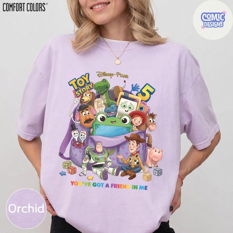 May include: A light purple t-shirt featuring a graphic print of Toy Story 5 characters including Woody, Buzz Lightyear, Rex, and Mr. Potato Head emerging from a purple backpack. The text reads Toy Story 5 and You've Got A Friend In Me.