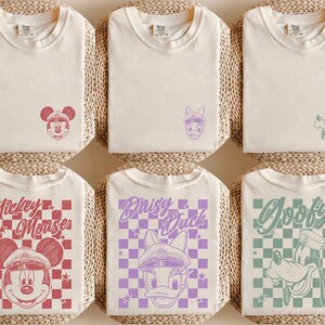 May include: Cream-coloured t-shirts with Mickey Mouse, Daisy Duck, and Goofy designs. The shirts feature graphics of Mickey Mouse in a red checkered pattern, Daisy Duck in a purple checkered pattern, and Goofy in a green checkered pattern.