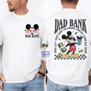May include: White t-shirt with a Mickey Mouse-inspired graphic. The front features the text "DAD BANK" with a Mickey Mouse head wearing sunglasses. The back displays Mickey with money and a coffee cup, with text "DAD BANK" and "OPEN 24/7 FAST CASH NO CREDIT CHECKS SAME DAY LOANS."