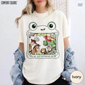 May include: An ivory short-sleeve t-shirt featuring a graphic of a frog-shaped frame containing various cartoon characters. The text "YOU'VE GOT A FRIEND IN ME" is printed below the characters. The shirt has a crew neckline and a relaxed fit.
