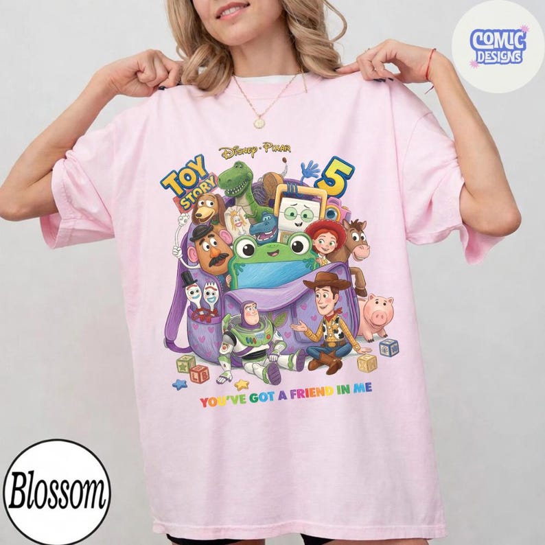 May include: A light pink t-shirt featuring a colourful graphic of Toy Story characters including Woody, Buzz Lightyear, Rex, and Mr. Potato Head emerging from a purple backpack. The text reads Toy Story 5 and You've Got A Friend In Me.