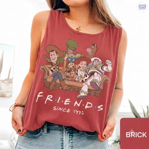 May include: A brick red sleeveless tank top featuring a graphic print of various animated characters sitting on a couch. Below the characters, the text reads "F R I E N D S" in white letters with colorful dots between them, followed by the text "SINCE 1995".