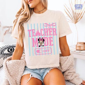 May include: Cream-colored t-shirt with the text "TEACHER MODE OFF" in pink and blue, featuring a Minnie Mouse graphic. The design includes a bow and vertical stripes. The shirt is short-sleeved.