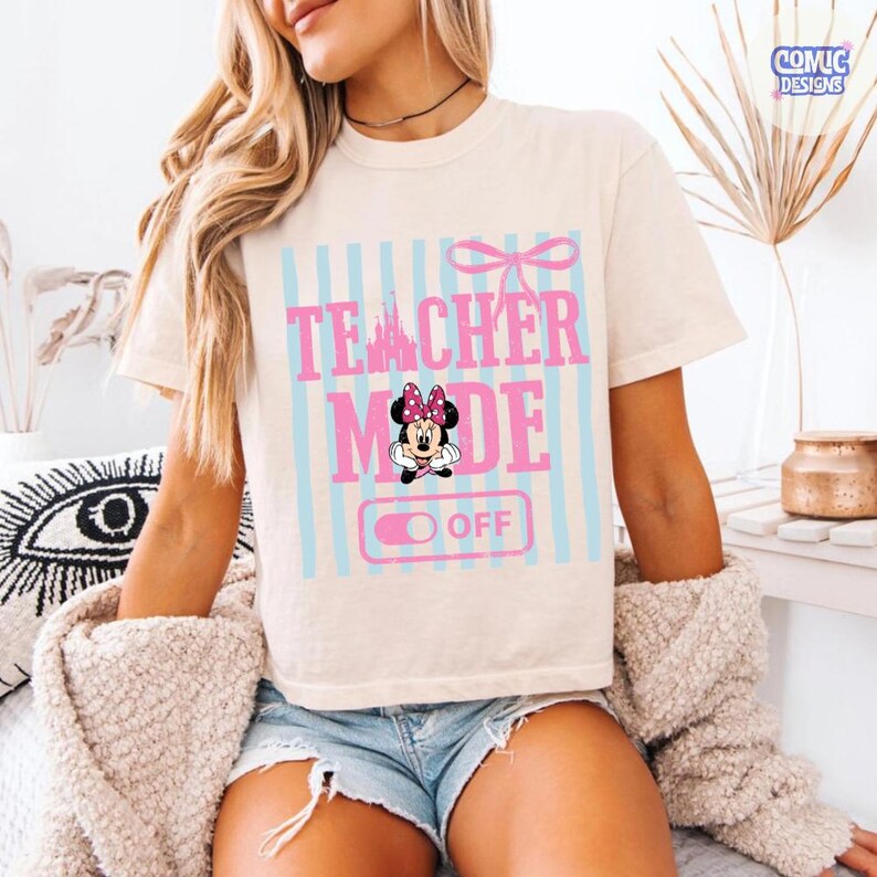 May include: Cream-colored t-shirt with the text "TEACHER MODE OFF" in pink and blue, featuring a Minnie Mouse graphic. The design includes a bow and vertical stripes. The shirt is short-sleeved.
