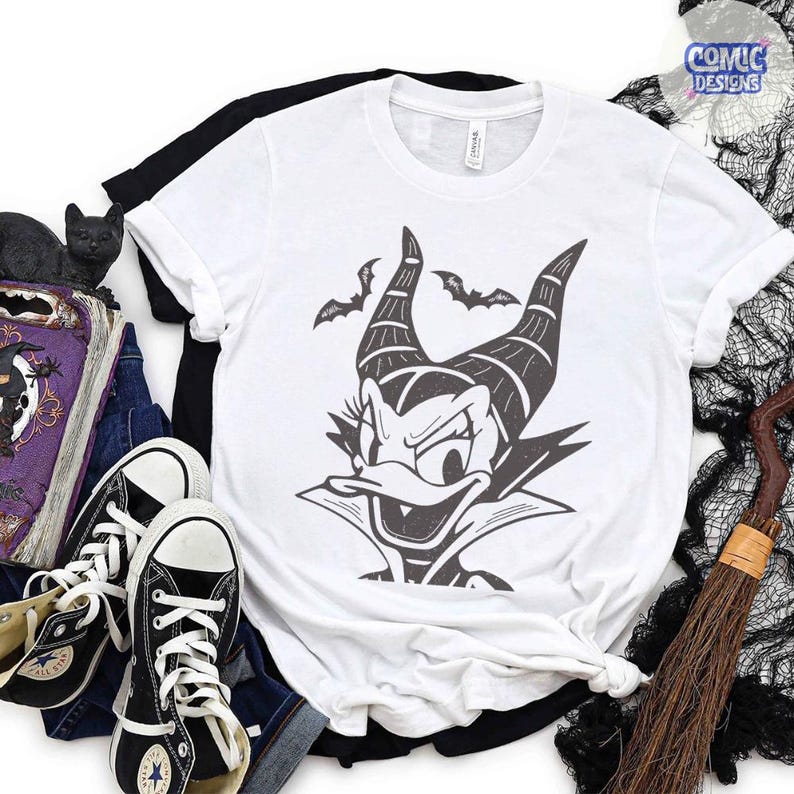 May include: White t-shirt featuring a black and gray graphic of a cartoon character with horns and bat silhouettes. The shirt is folded with a black t-shirt underneath, paired with black sneakers and a witch's broom. The words "Comic Designs" are in the upper right corner.