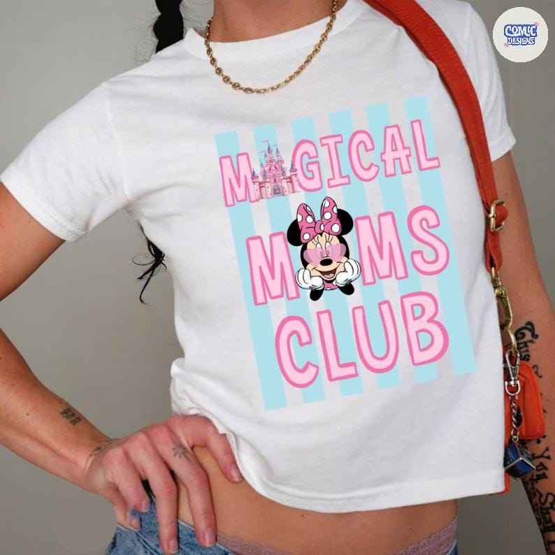 K&ouml;nnte beinhalten: Wei&szlig;es T-Shirt mit dem Text "MAGICAL MOMS CLUB" in Rosa, mit einer Minnie-Maus-Grafik und einem rosa Schloss. Der Text befindet sich vor einem Hintergrund aus hellblauen vertikalen Streifen. Eine goldene Halskette und eine rote Umh&auml;ngetasche sind ebenfalls zu sehen.